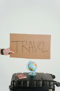 valigia nera con sopra piccolo mappamondo e passaporto. Una mano che compare da sinistra e tiene un cartone con su scritto Travel. Viaggi economici giugno 2026